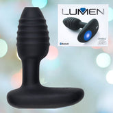 A black, ribbed-texture plug-shaped device with a flat base is displayed against a softly blurred light background. The inset image reveals the product name "OhMiBod Lumen Vibrating Butt Plug" alongside its boxed version, labeled as Bluetooth enabled by Kiiro and OhMiBod. With LED Brilliance, this vibrating butt plug offers an immersive experience.
